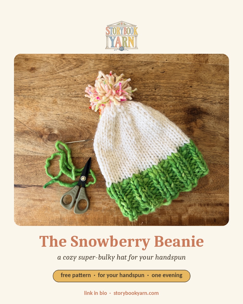 The Snowberry Beanie — a snowy white super-bulky beanie with a green handspun brim and a pompom, on a wood backdrop.